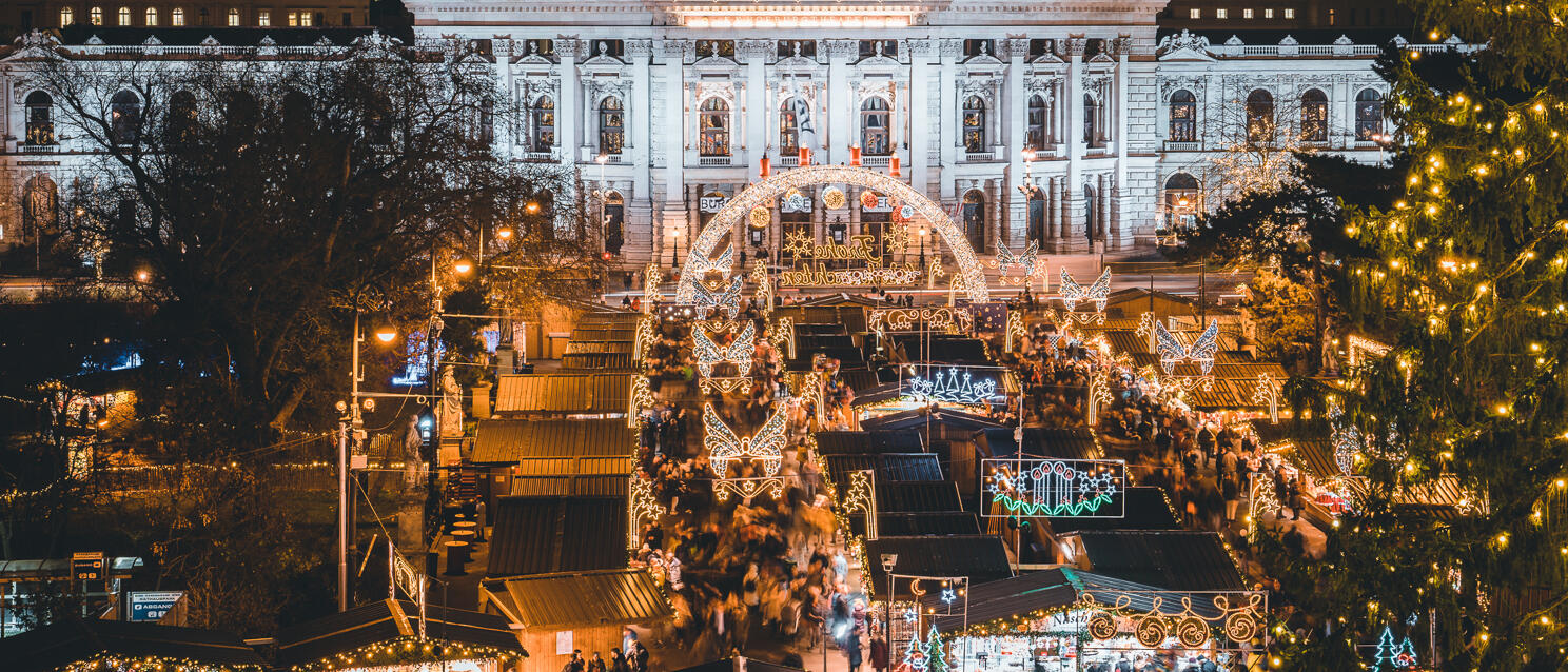 Vienna Christmas market at City Hall Square at night, illuminated stalls and light arches, Burgtheater in background.
