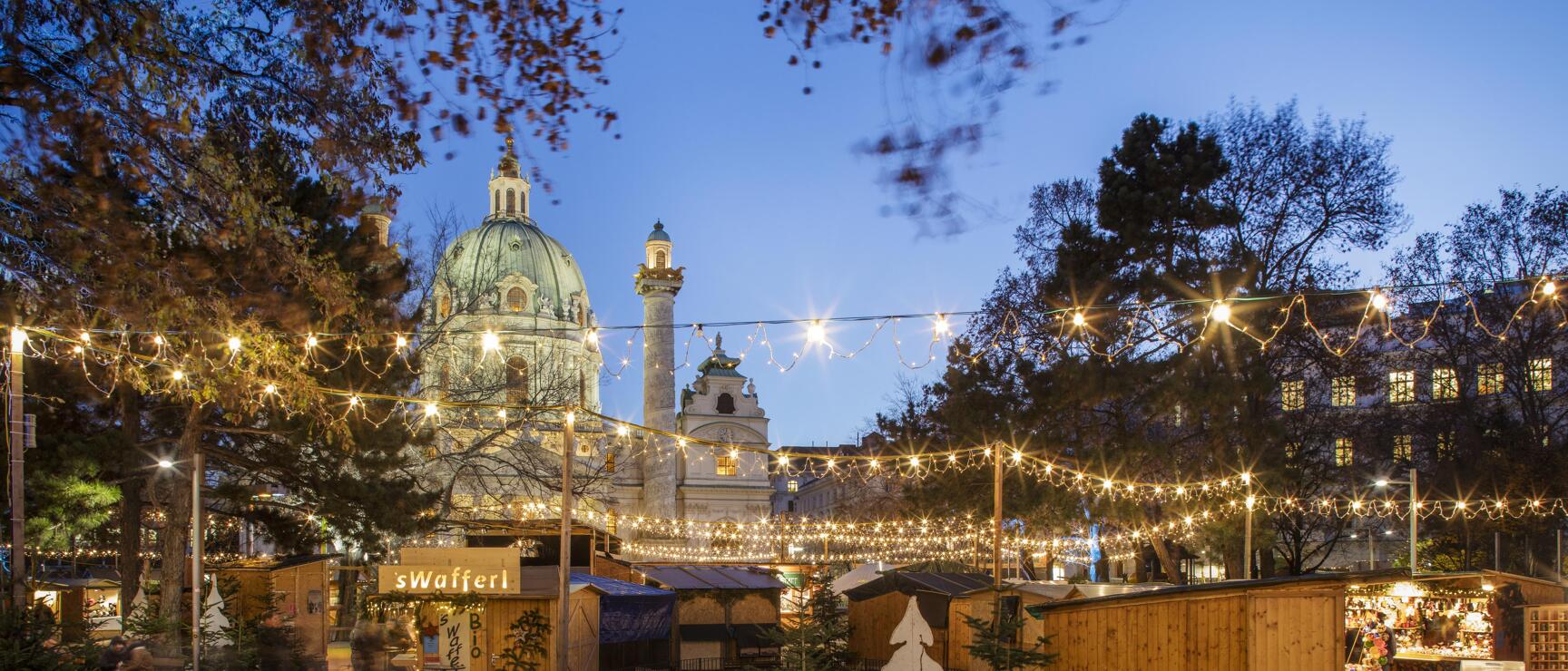 Christmas market at Karlsplatz Vienna in evening light, wooden huts with fairy lights, Karlskirche in the background.