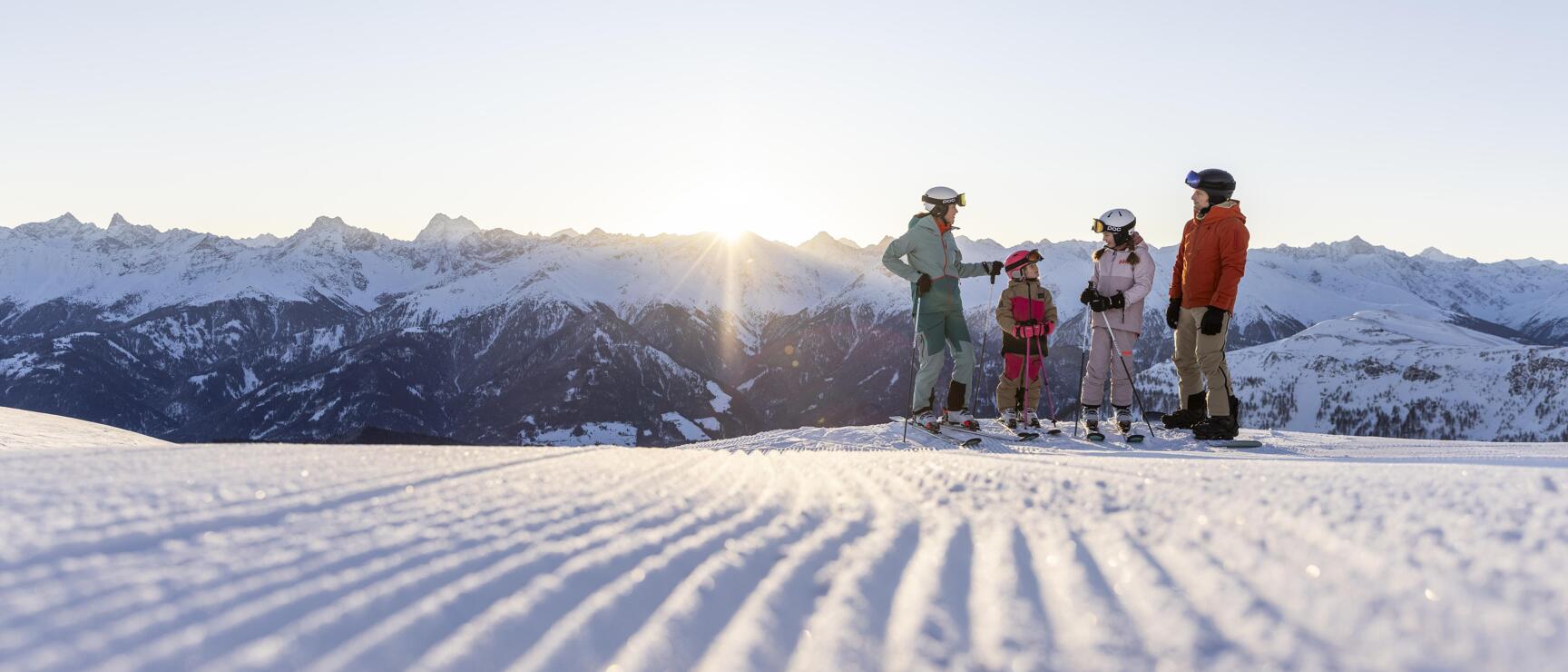 Four skiers on prepared slope at sunrise, snow-covered mountain range in background.