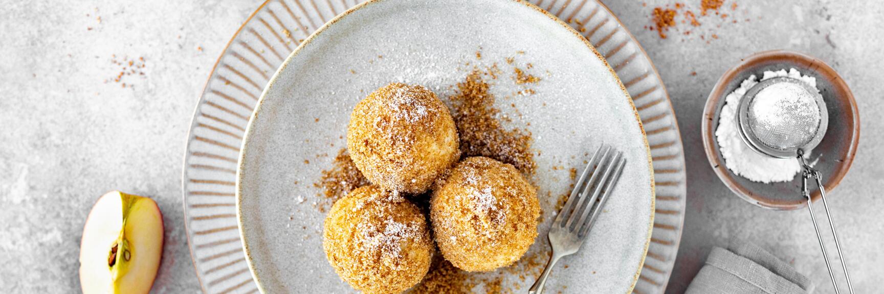 Three apple dumplings with breadcrumbs and icing sugar on grey plate, halved apples and sugar sifter beside.