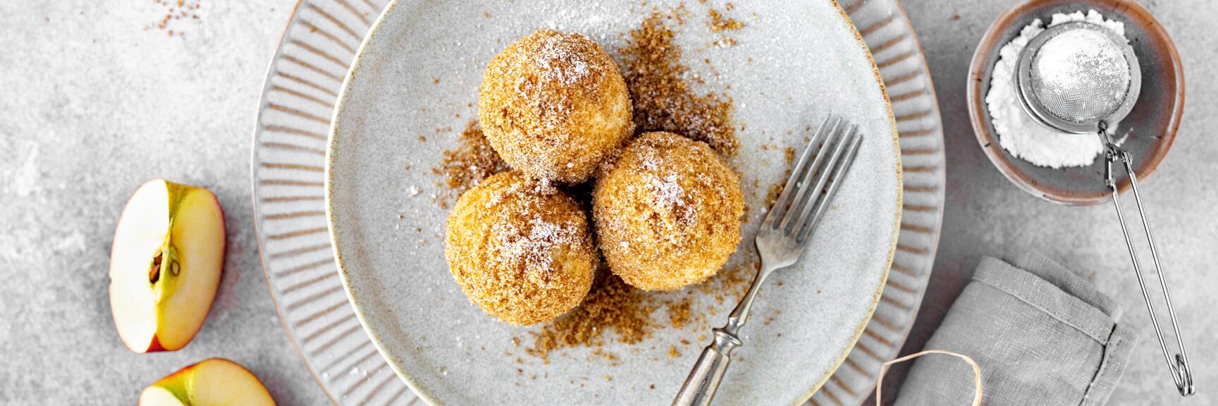 Three apple dumplings with breadcrumbs and icing sugar on grey plate, halved apples and sugar sifter beside.