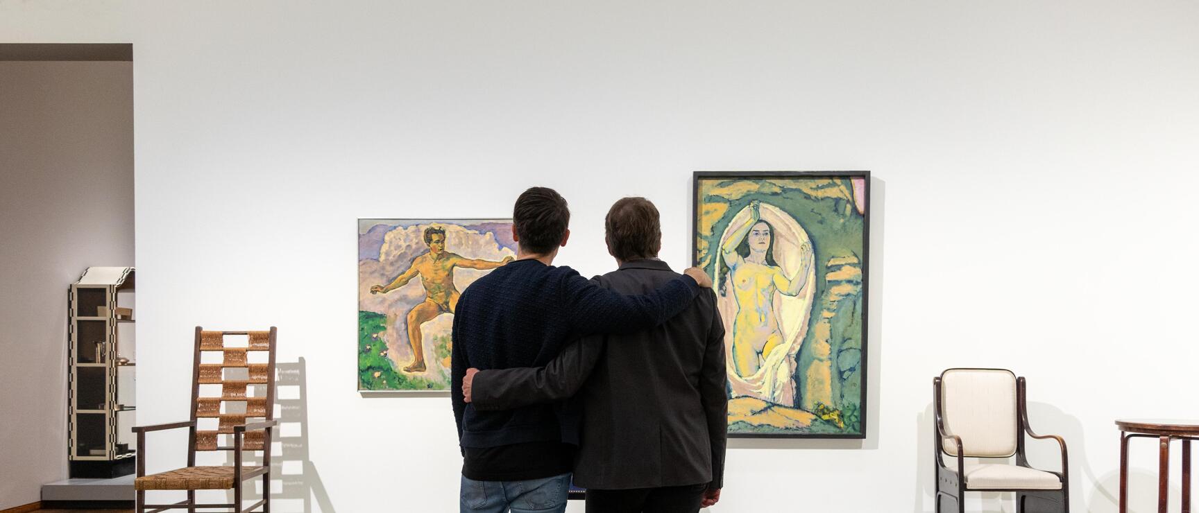 Two men with arm around shoulder viewing a painting in a museum, antique chairs positioned to the side.