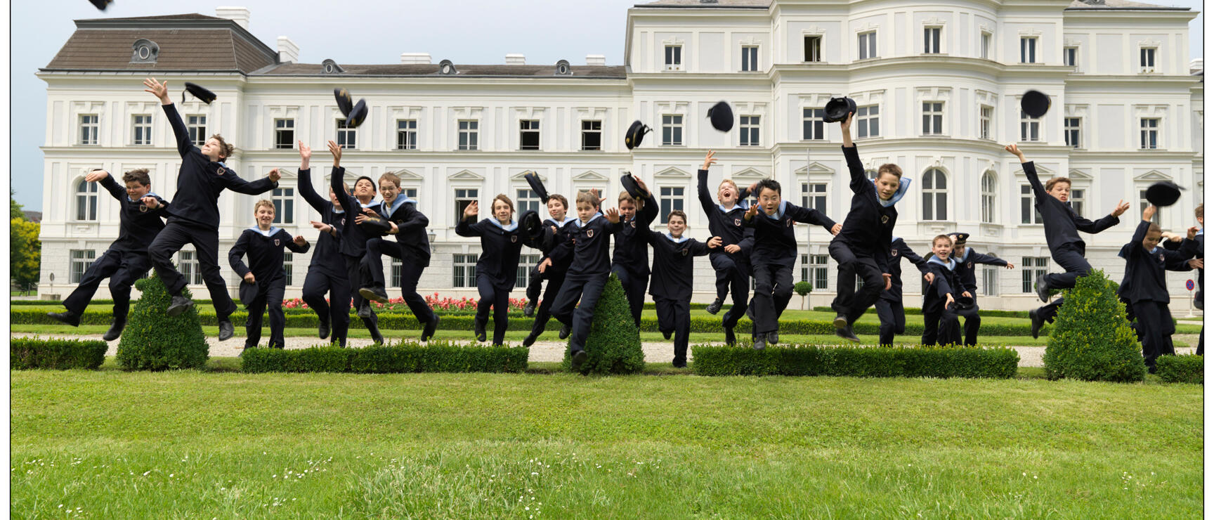 Boys in black uniforms jumping in front of white castle, tossing caps in the air, manicured lawn.
