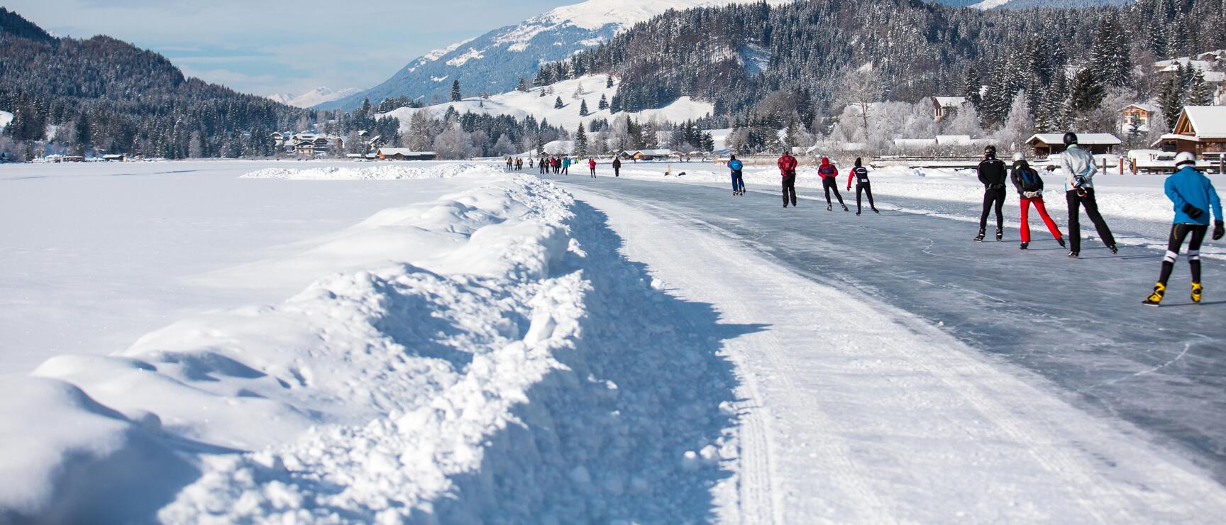 Multiple people on a natural ice rink, snow-covered mountains and coniferous forests in background.