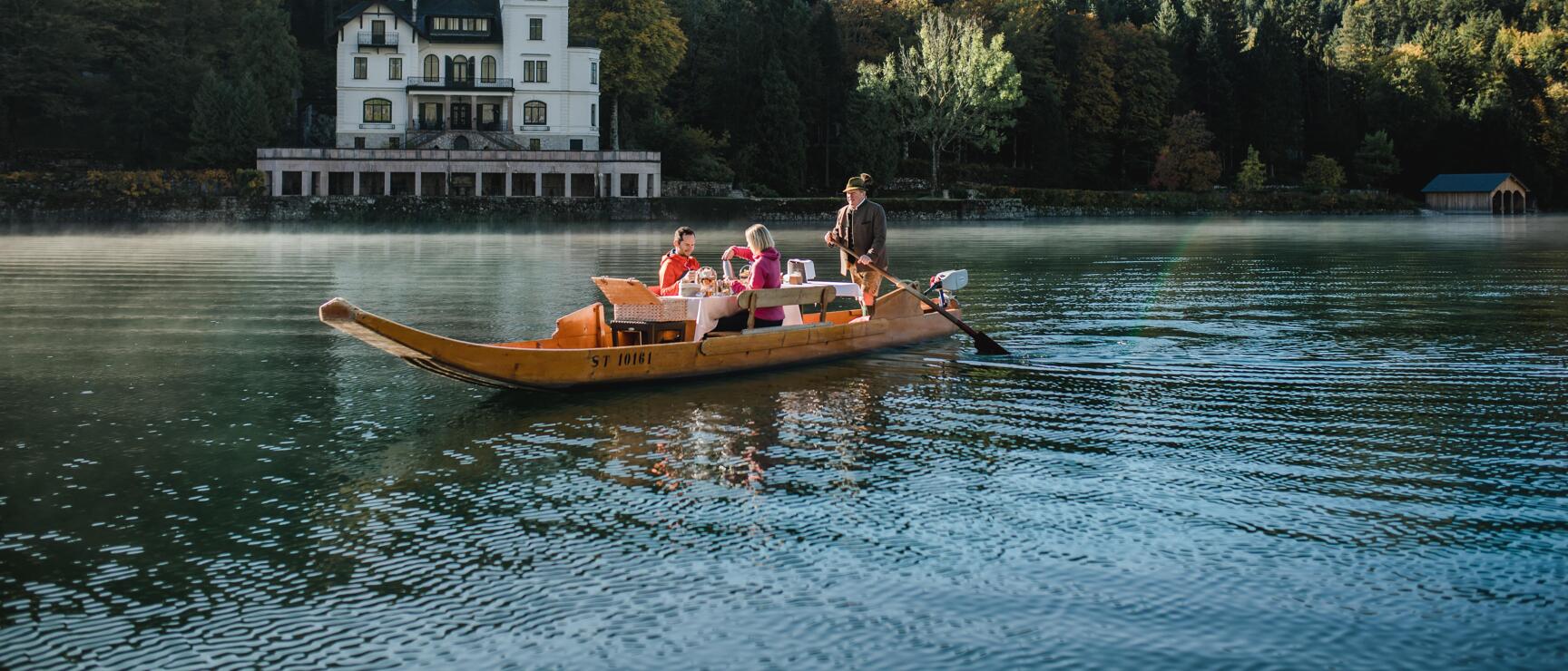 Wooden boat with three people on a lake, white villa and autumn forest in the background.