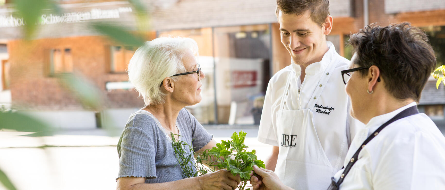 Two chefs in JRE aprons receiving herb basket from elderly woman, wooden building in the background.