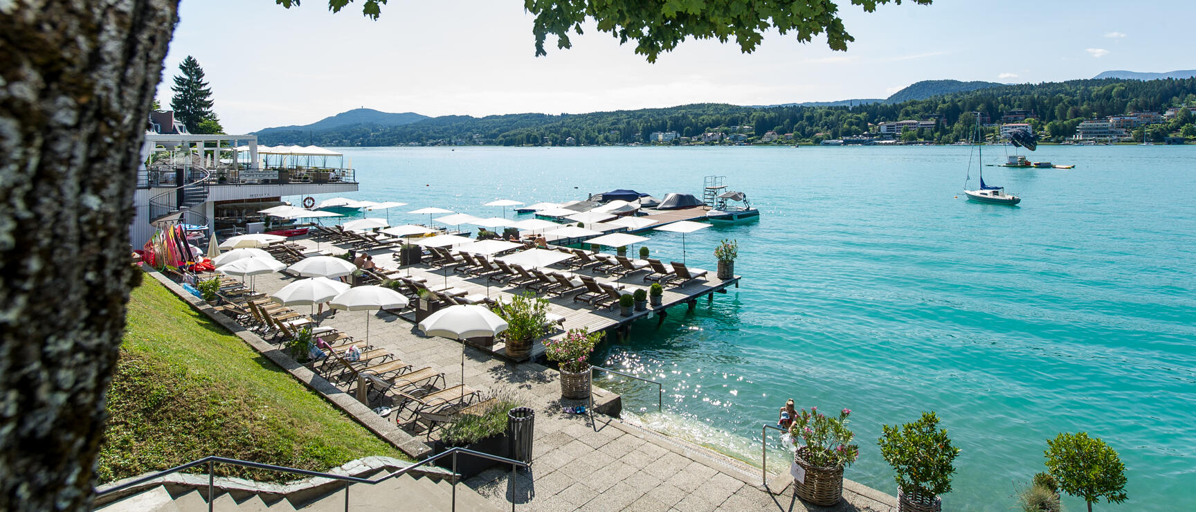Beach area with loungers and white parasols at a turquoise lake, mountains in the background.