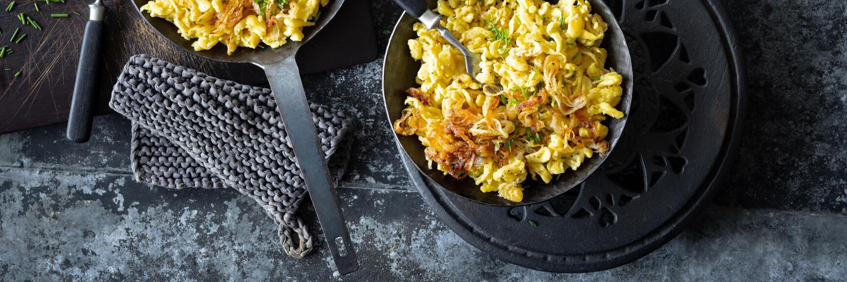 Two pans with cheese spätzle, crispy onions and chives, mortar and fork on dark surface.