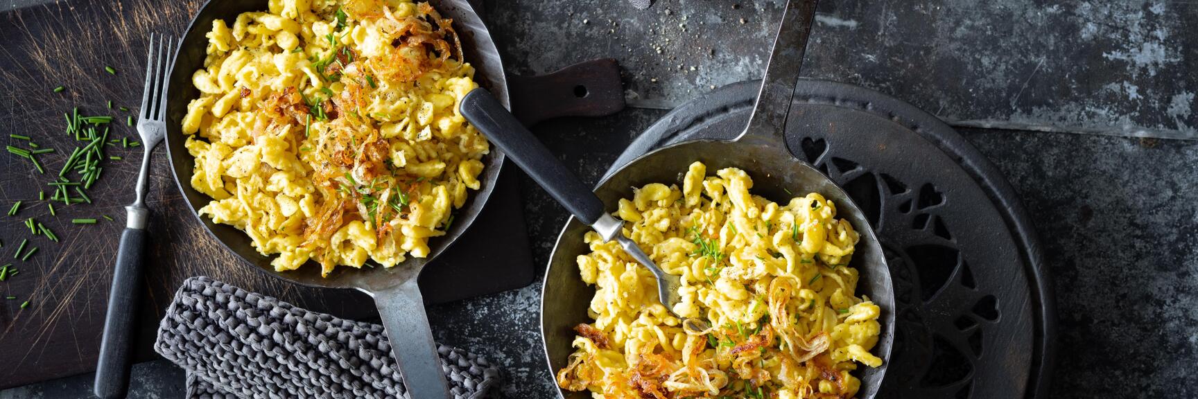Two pans with cheese spätzle, crispy onions and chives, mortar and fork on dark surface.