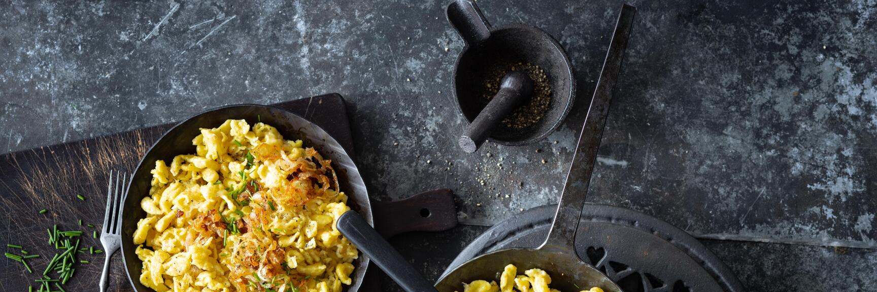 Two pans with cheese spätzle, crispy onions and chives, mortar and fork on dark surface.