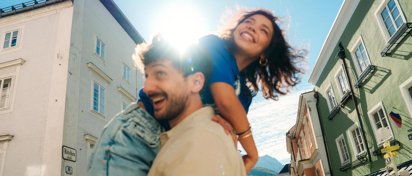 Woman laughing sitting on a man's shoulders in an old town alley, lake and mountains in the background.