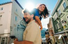 Woman laughing sitting on a man's shoulders in an old town alley, lake and mountains in the background.