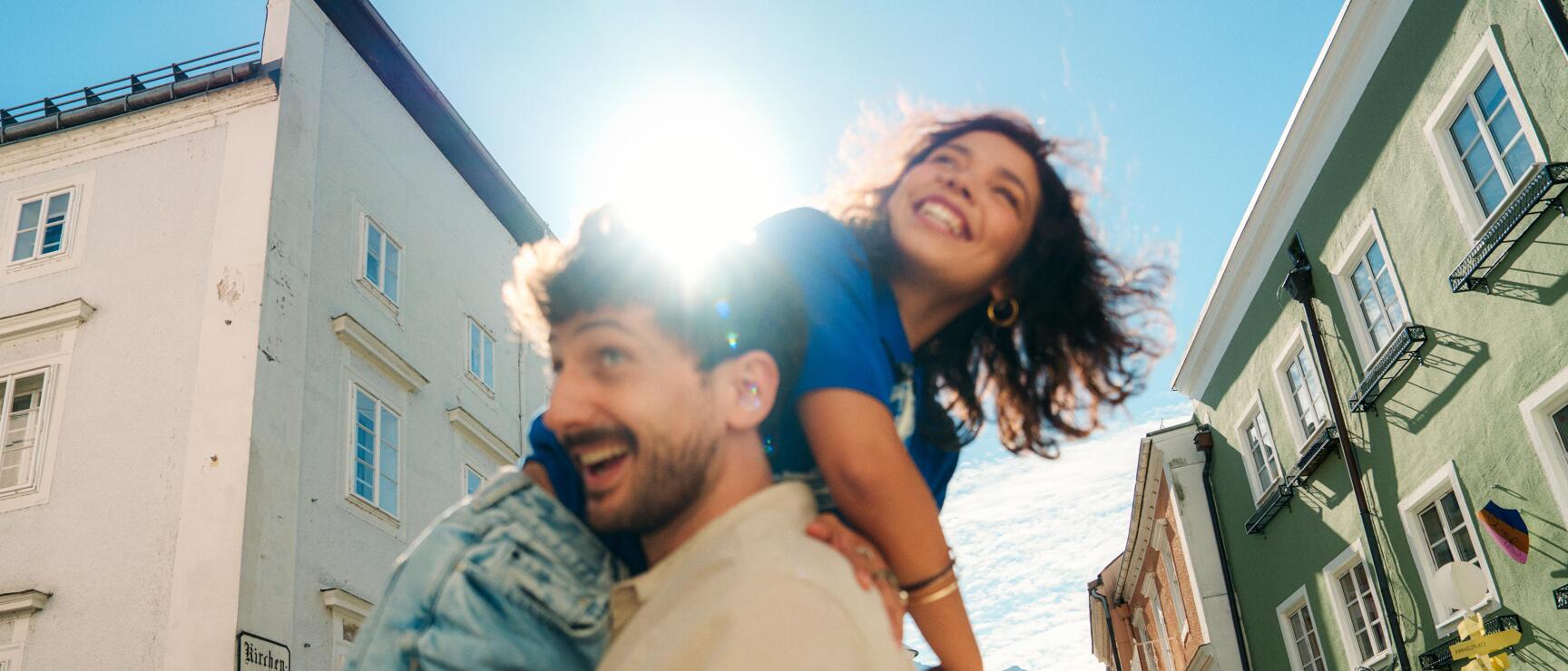 Woman laughing sitting on a man's shoulders in an old town alley, lake and mountains in the background.