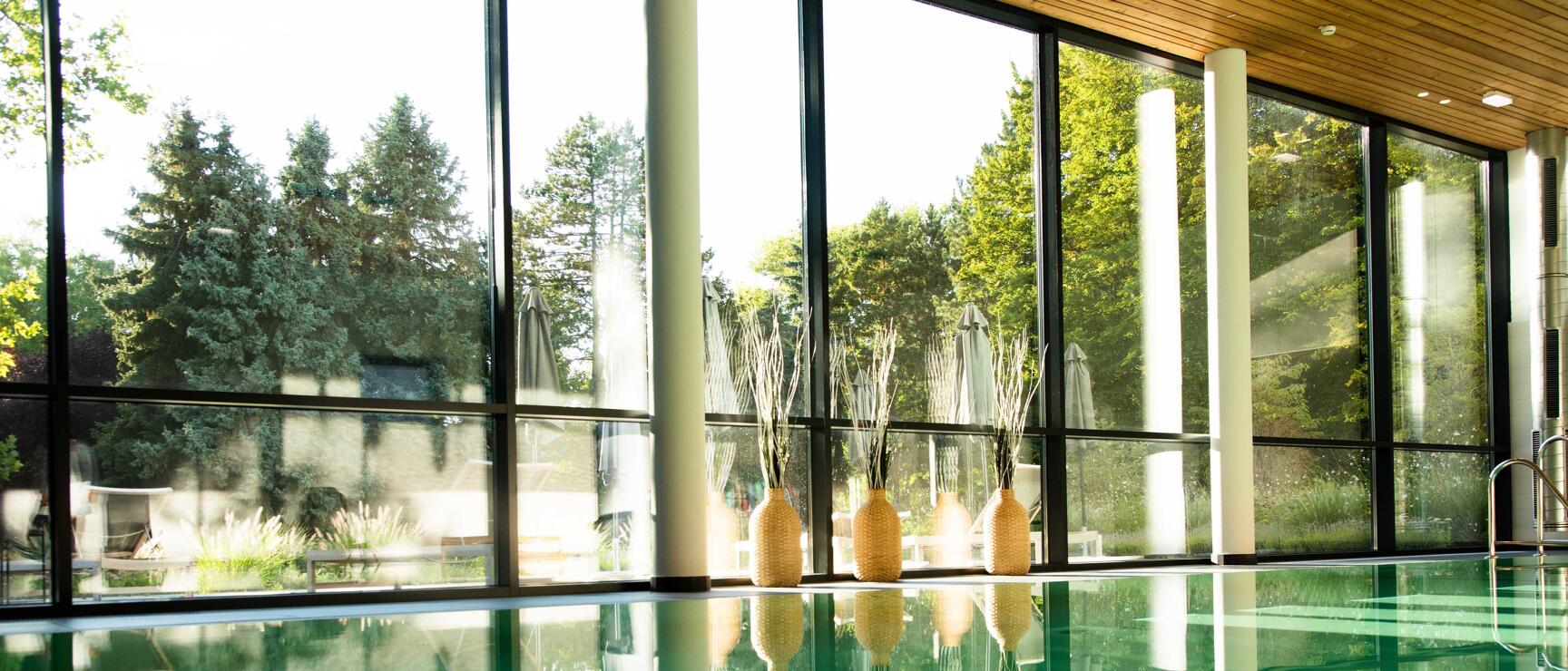 Indoor pool with green water, wicker baskets at pool edge, large glass front overlooking trees.