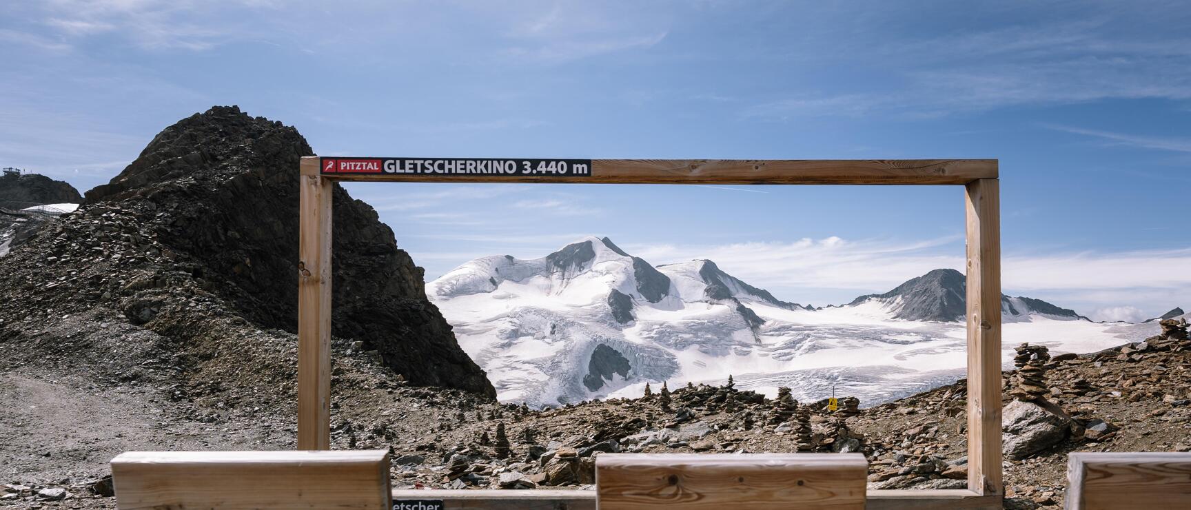 Wooden frame 'Pitztal Glacier Cinema 3,440 m' with glacier view and wooden benches in the foreground.
