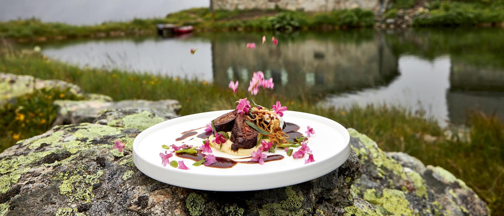 Plated meat dish with pink blossoms on white plate, rock, alpine lake and stone house in background.