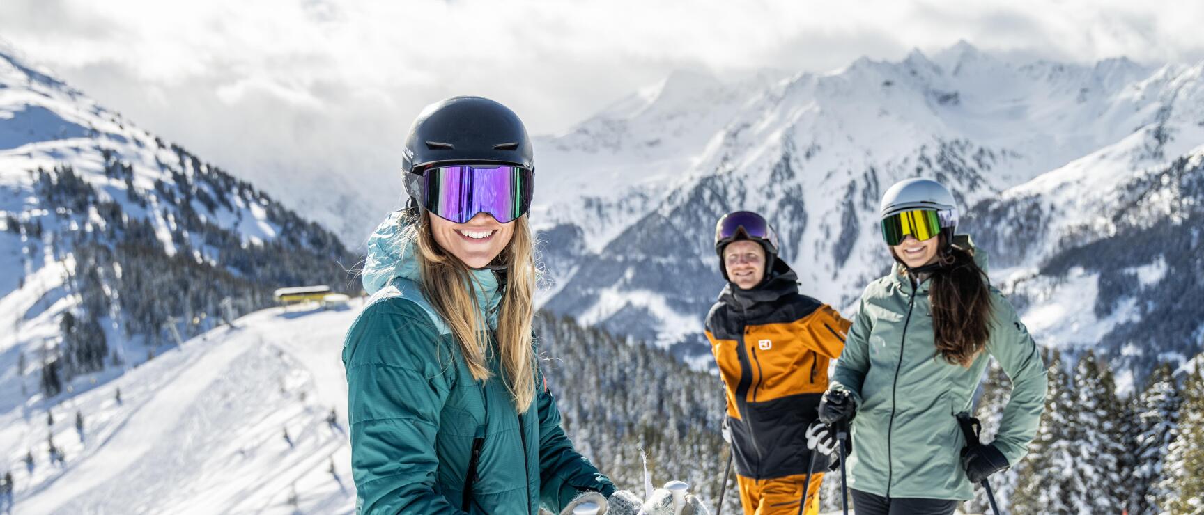 Three skiers wearing helmets and ski goggles on a slope with snow-capped Alps in the background.