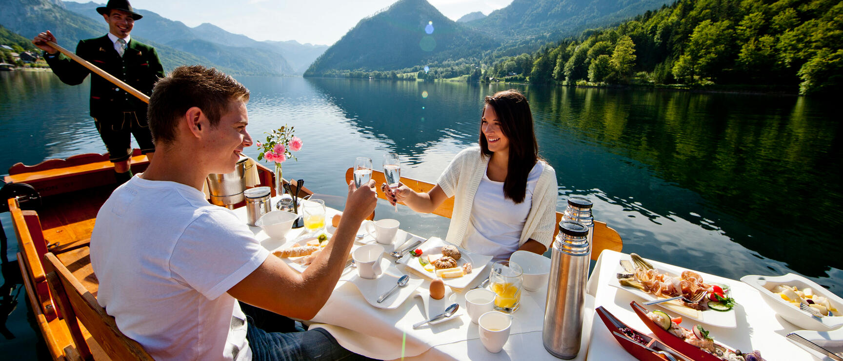 Couple toasting with glasses on a wooden boat, set breakfast table, gondola operator, mountain lake in the background.
