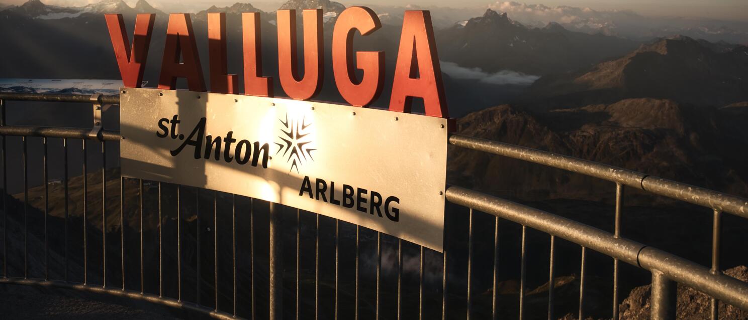 Sign reading 'Valluga – St. Anton Arlberg' on summit platform, vast Alpine panorama in evening light behind.