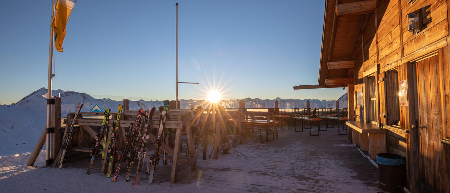 Ski lodge at sunset with ski racks, wooden terrace with benches, snow-covered mountains in background