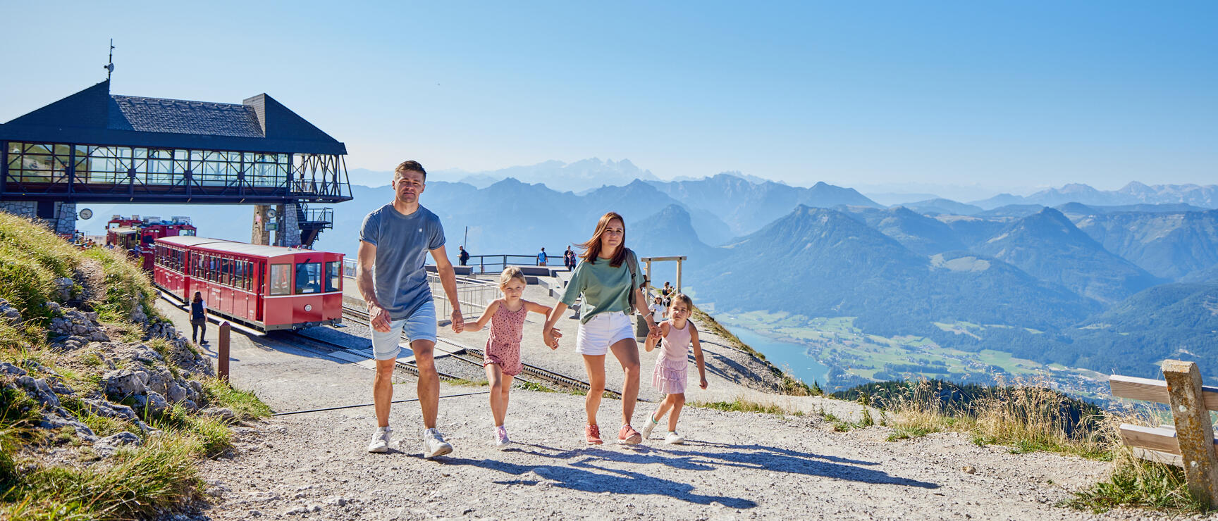 Two adults and two children on gravel path at Schafberg, red cog railway and alpine panorama in background