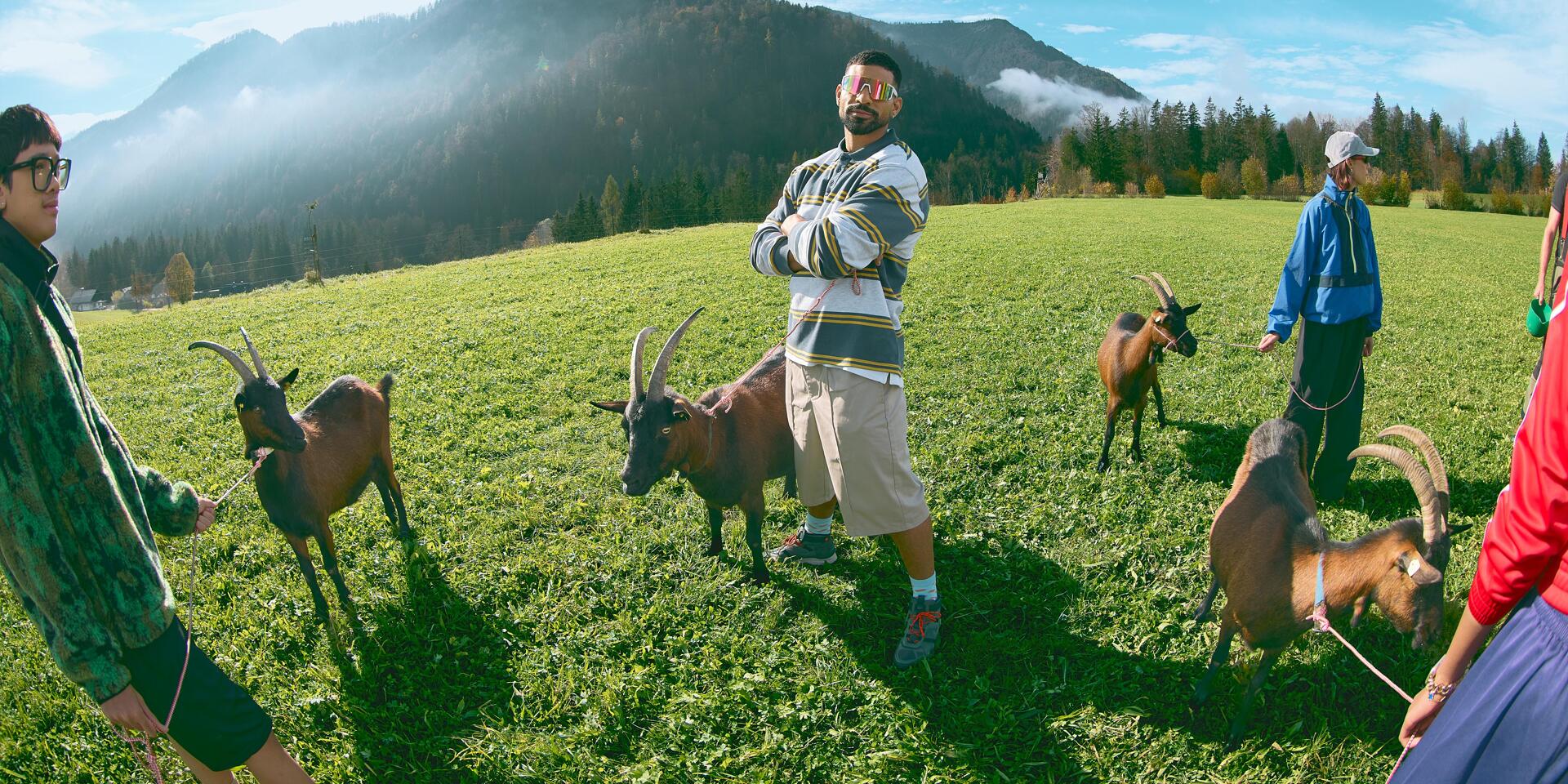 Three people with goats on an alpine meadow, forested mountain in the background.