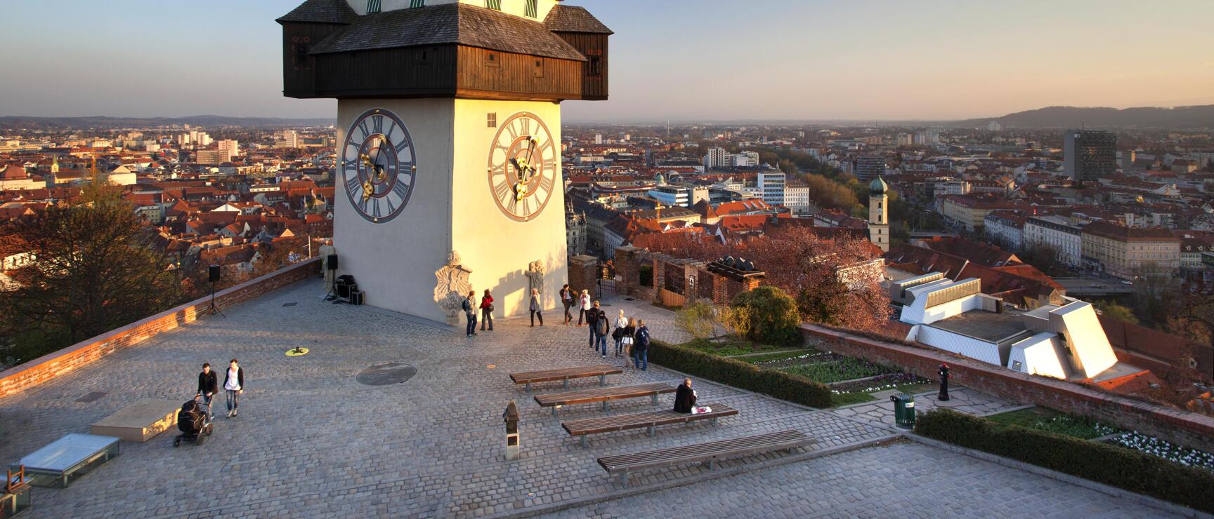 Clock tower on Schlossberg in Graz at sunset light, city panorama in background, visitors on the square.