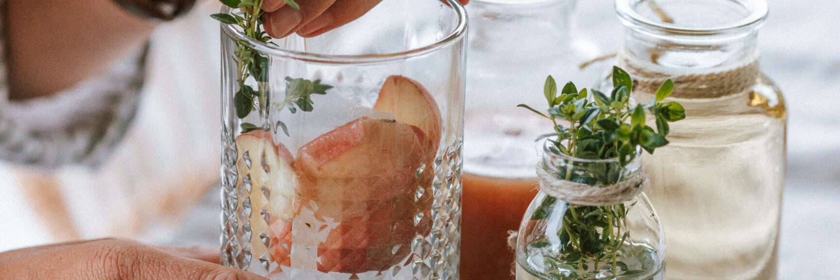 Hands placing herb branch in glass with peach pieces, small herb bottles beside on tray.