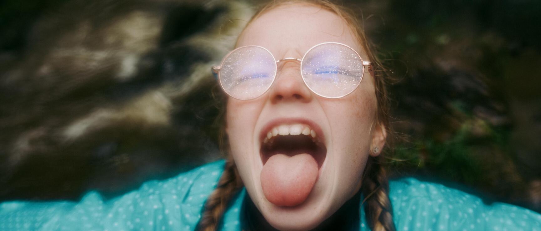 Girl with braids, round glasses and turquoise jacket sticking out her tongue, blurred background.