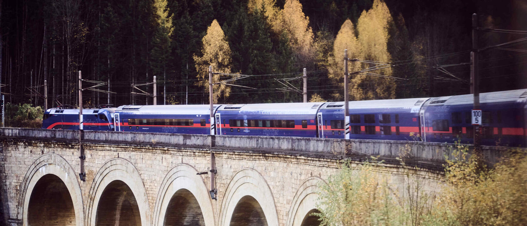 ÖBB train on a stone viaduct with arches over a river, autumn forest in the background.