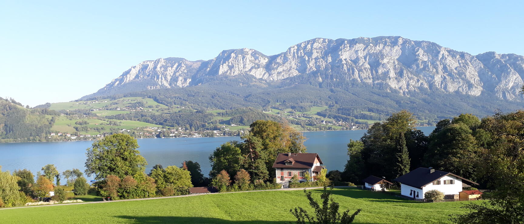 Green meadows with two houses by Lake Atter, trees, with Höllengebirge rock formations in the background.