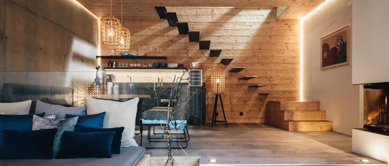 Modern chalet interior with wood walls, sofa with blue cushions, staircase and fireplace with warm lighting.