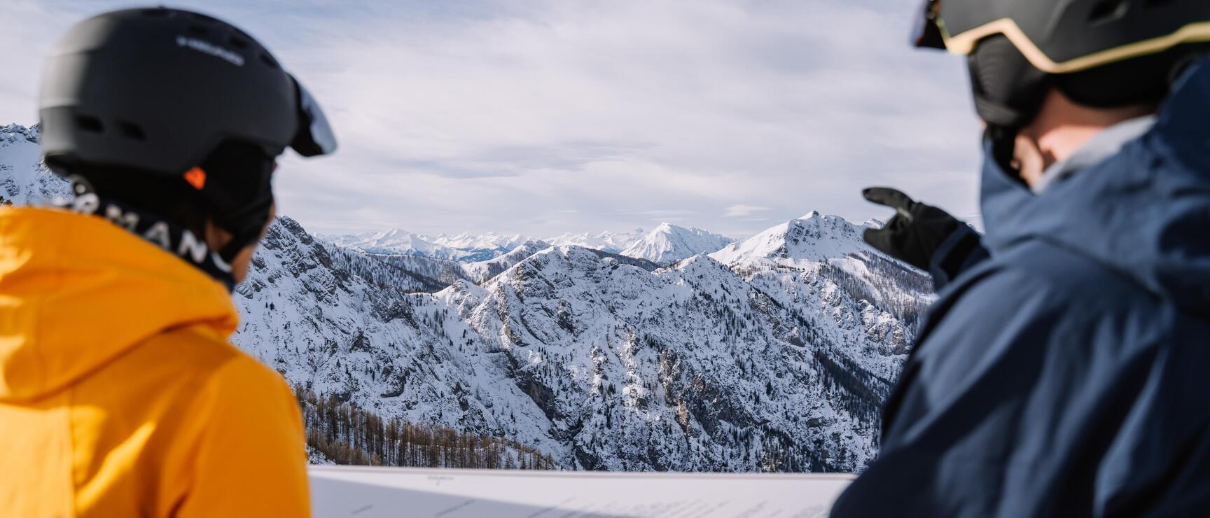 Two skiers with helmets looking at snow-capped mountain peaks, information board in foreground.