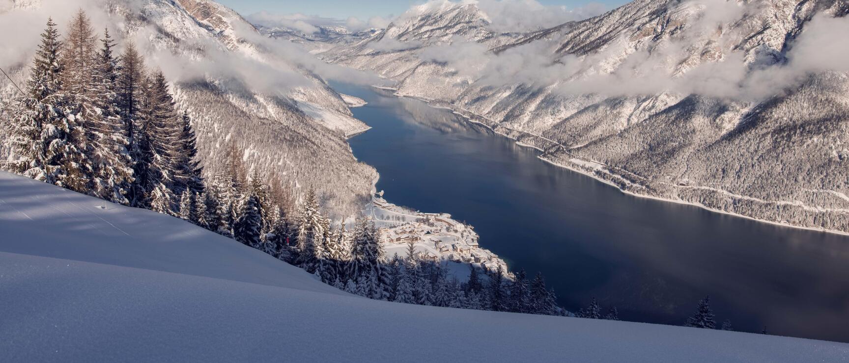 Winter view of Lake Achensee with snowy mountains and forests, small village on the shore, blue sky.