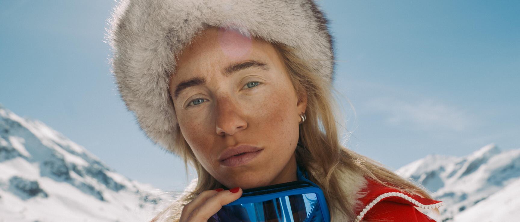 Woman wearing white fur hat, red ski jacket and blue ski goggles in front of snow-covered mountains.