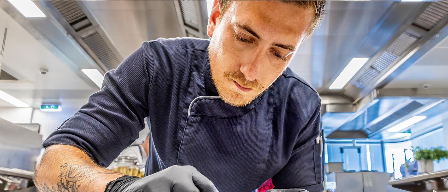 Chef in black gloves garnishing a dish on dark plate in professional kitchen.