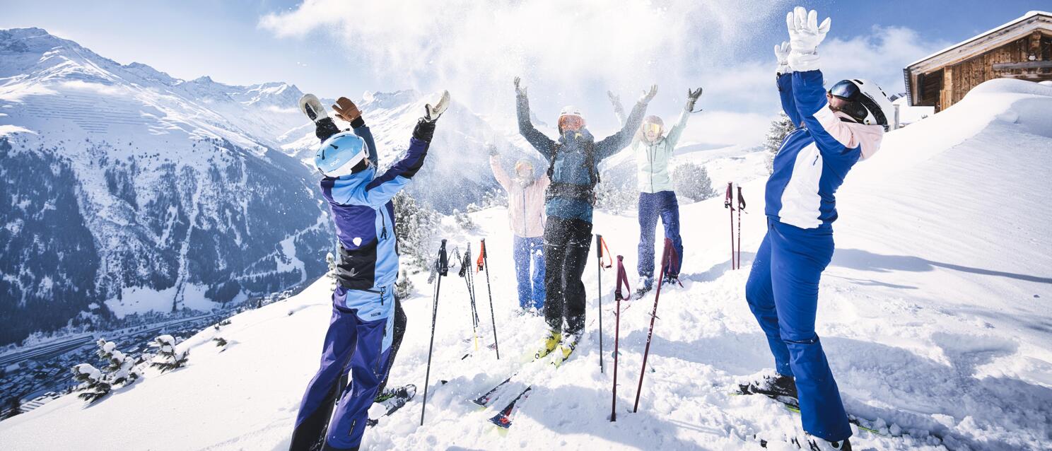 Group of skiers throwing snow in the air, mountain panorama and alpine hut in the background.