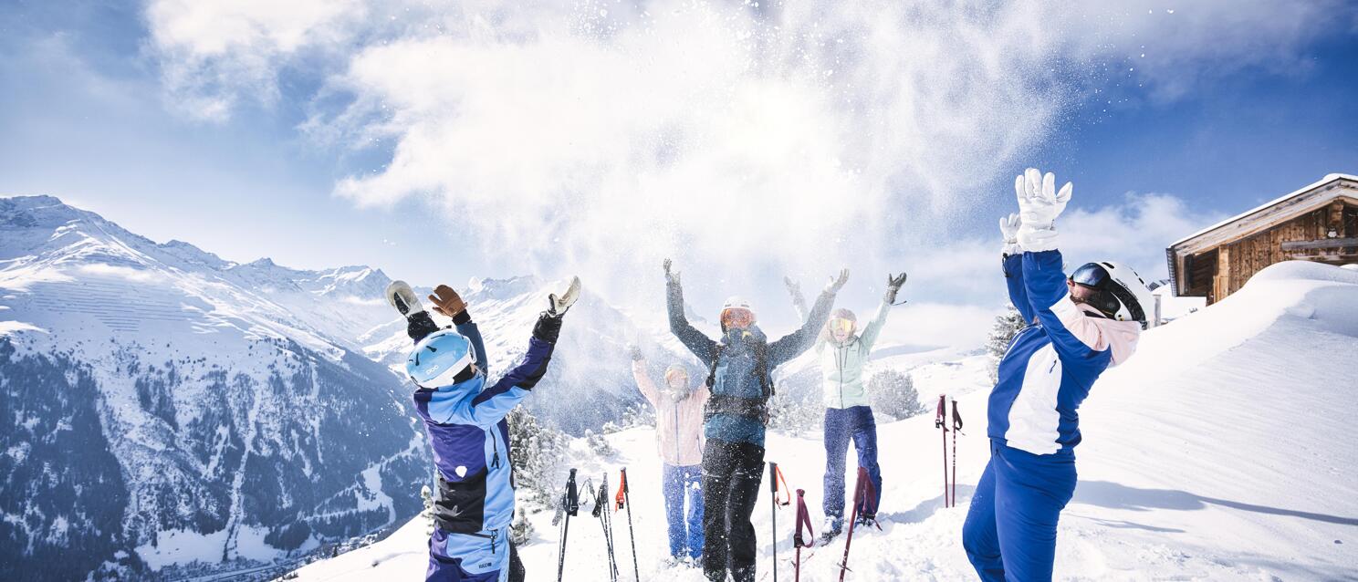 Group of skiers throwing snow in the air, mountain panorama and alpine hut in the background.