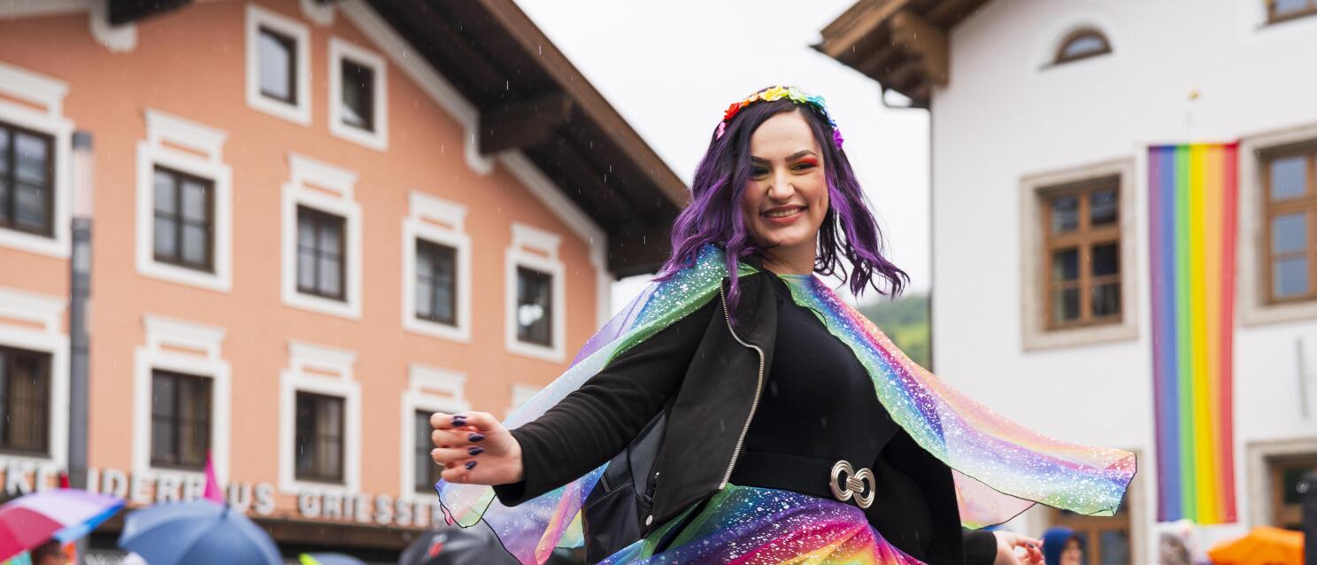 Eine Person in einem regenbogenfarbenen Outfit mit Umhang dreht sich fröhlich bei einer Pride-Veranstaltung in Mittersill.