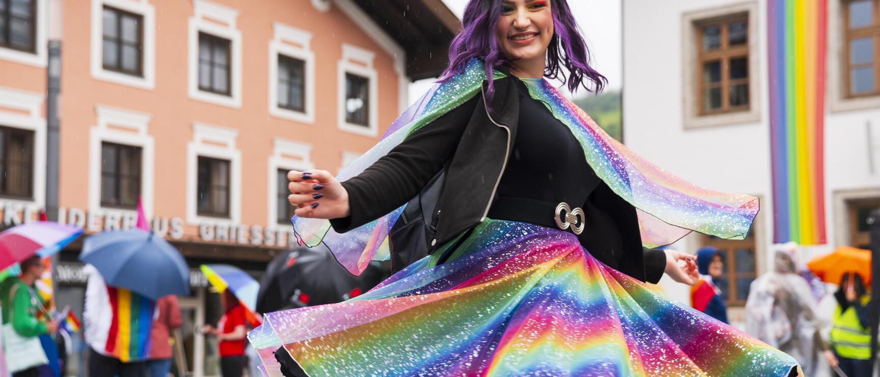 Person wearing a rainbow-colored outfit and cape twirls joyfully during a Pride event in Mittersill, with pastel buildings, umbrellas, and a rainbow flag in the background on a rainy day.
