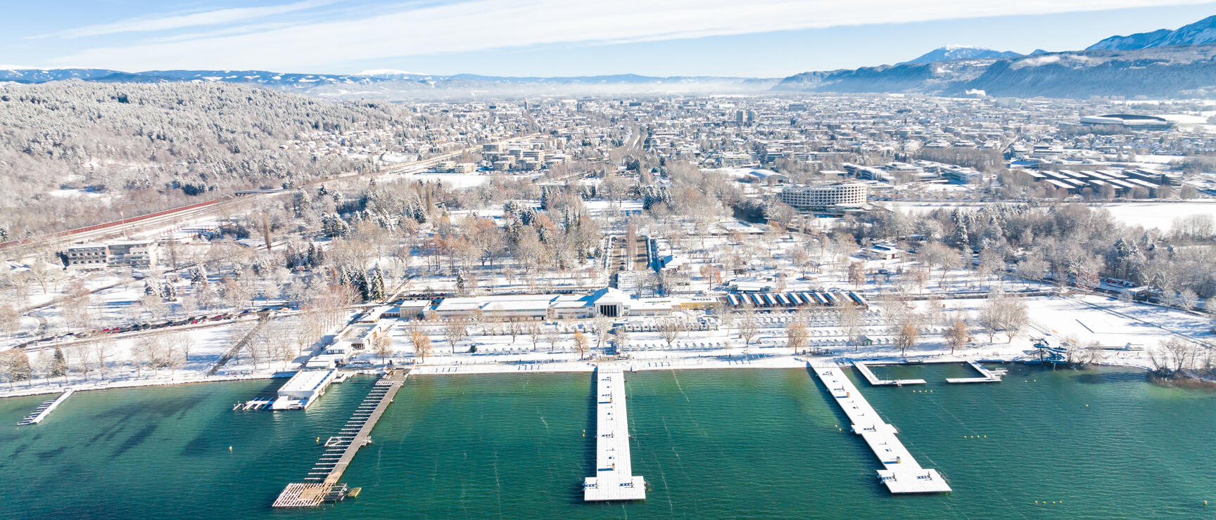 Aerial view: snow-covered lakeside with bathing jetties in winter, city and mountains in background, Lake Constance.