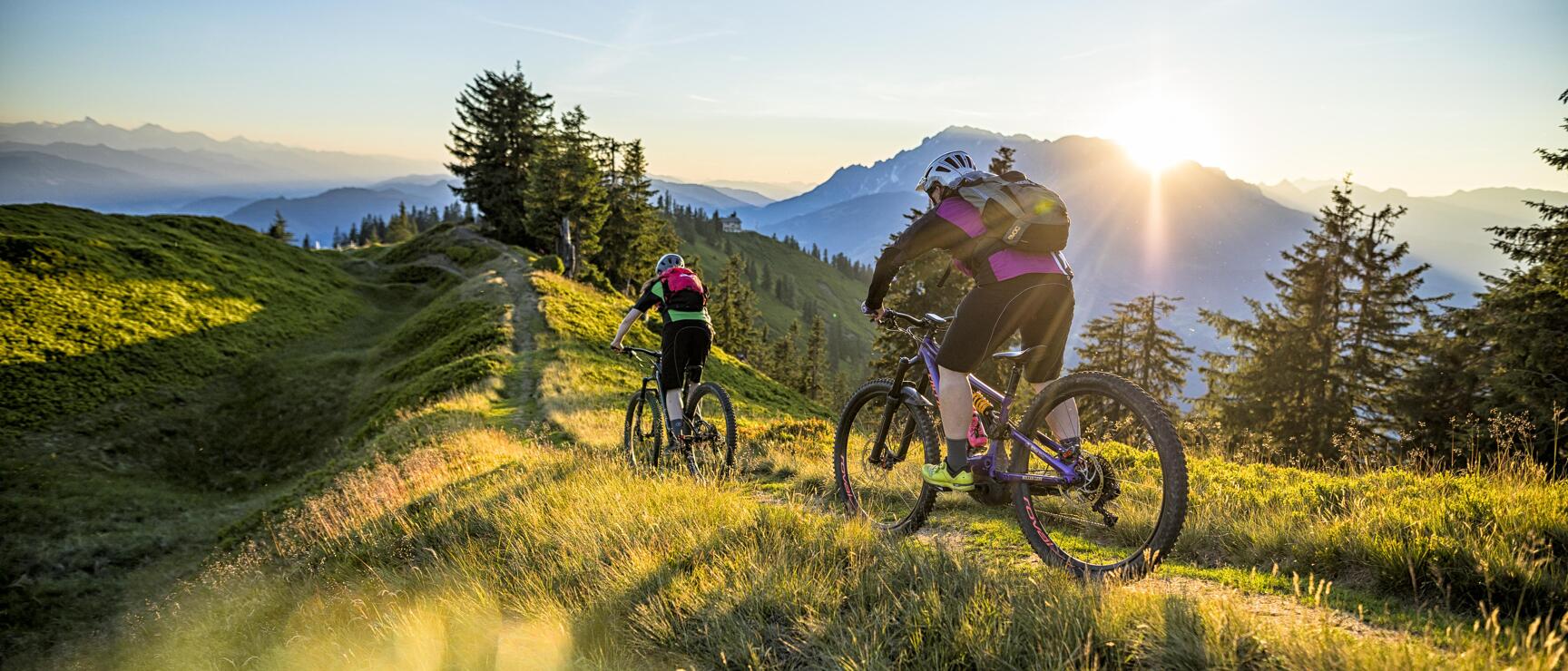 Two mountain bikers on a mountain trail at sunset with mountain panorama in the background.