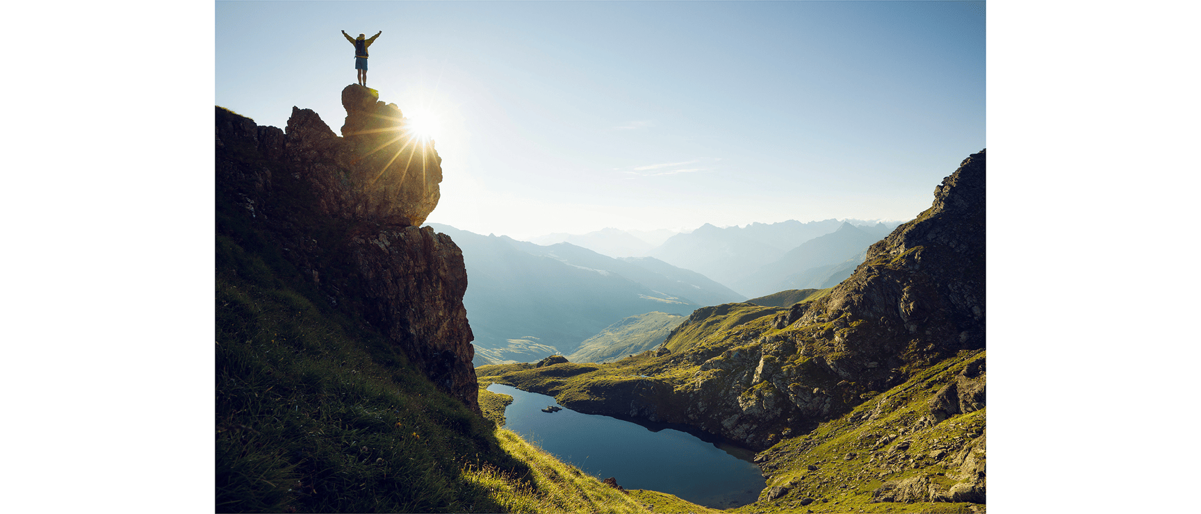 Person with backpack standing on rocky outcrop, arms outstretched, mountain lake and alpine panorama in background.