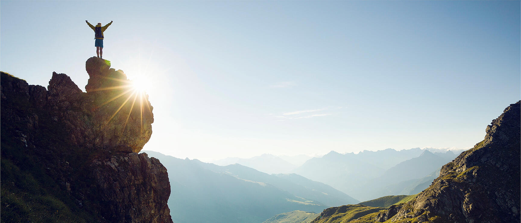 Person with backpack standing on rocky outcrop, arms outstretched, mountain lake and alpine panorama in background.