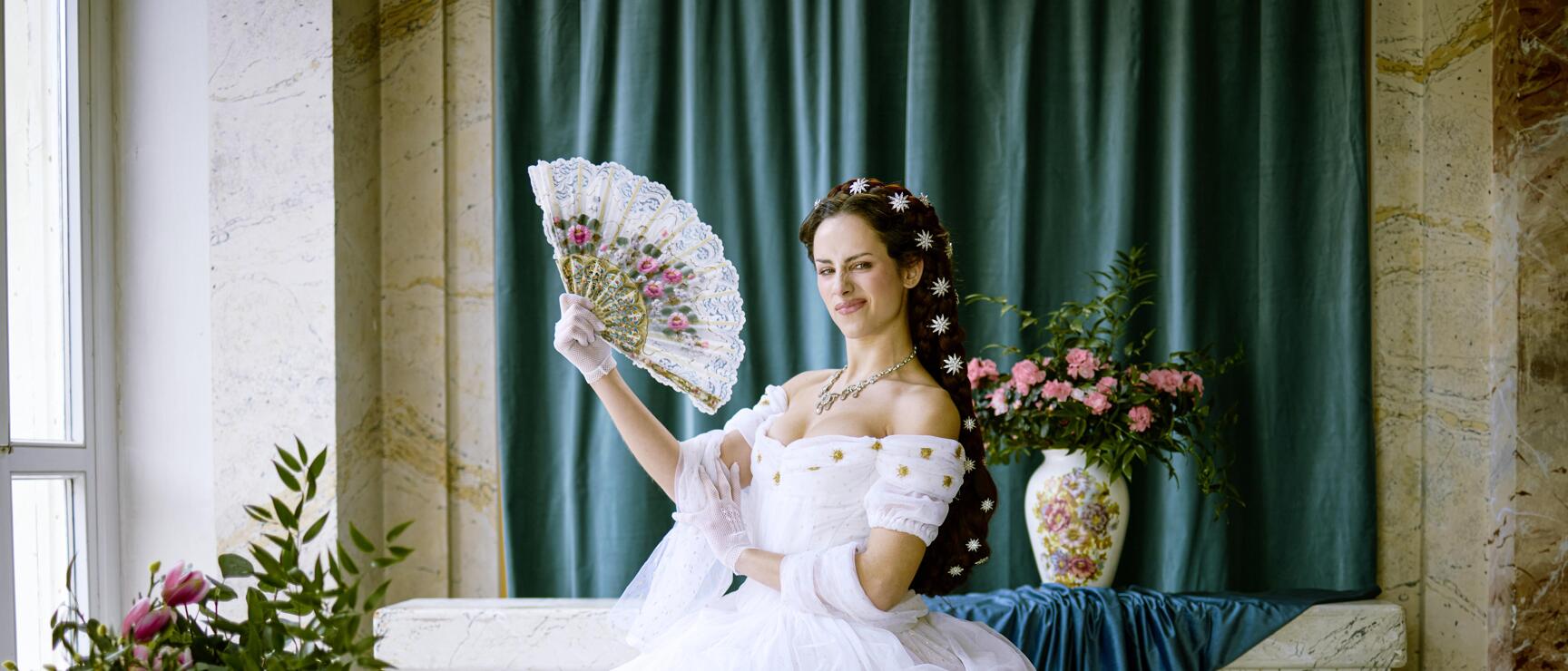 Woman in white ball gown with lace fan, hair styled updo with flowers, sitting in ornate marble hall.