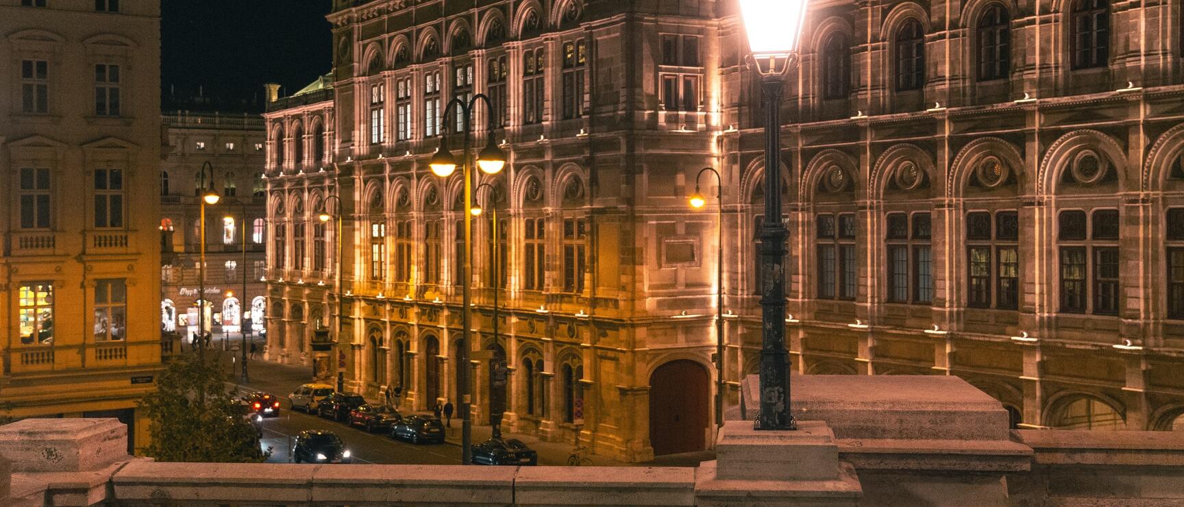 Vienna State Opera at night, illuminated, with street lamp in the foreground and parked cars on the street.