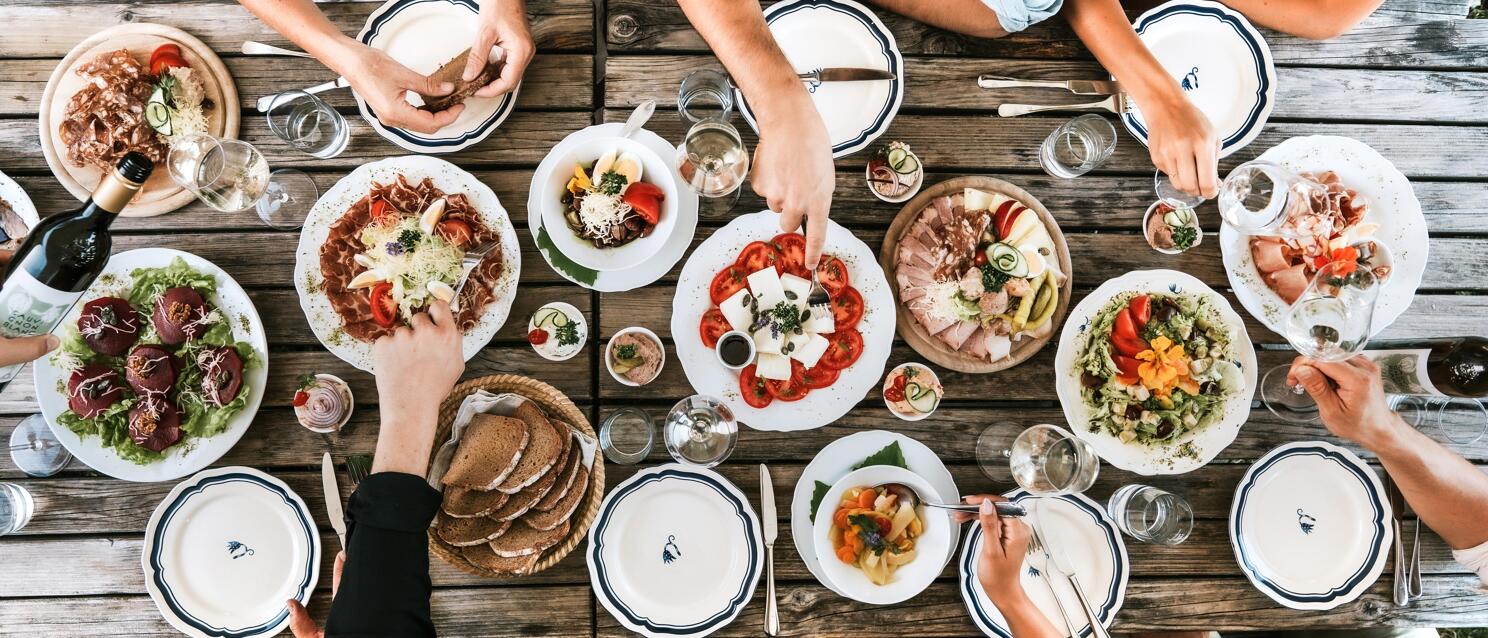 Bird's eye view of rustic wooden table with salads, cold cuts, bread and soups, hands reaching for food