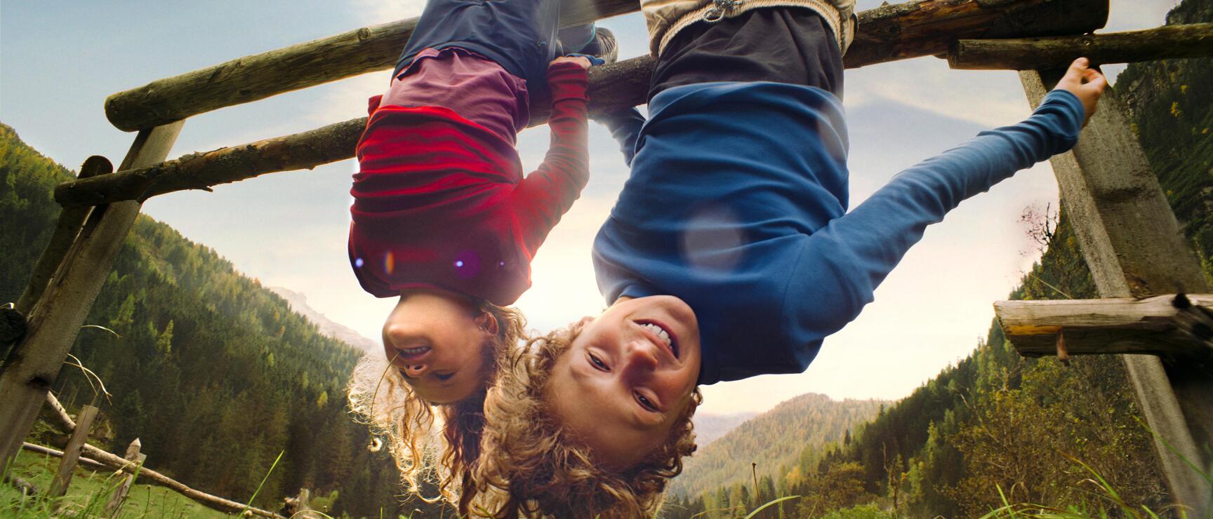 Two children hanging upside down laughing on a wooden fence, green alpine meadow and mountain forest in background.