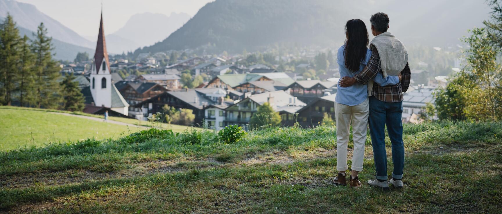Two people standing on a meadow overlooking an Alpine village with church tower and mountains in the background.