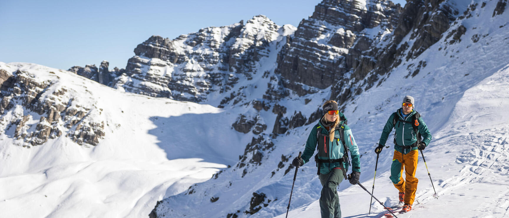 Two ski mountaineers ascending a snow-covered slope with rocky peaks and blue sky in the background.